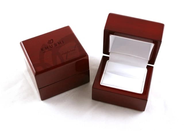 A glossy dark red-brown wooden ring box, open to show a white interior, with the Khushi logo embossed on the lid