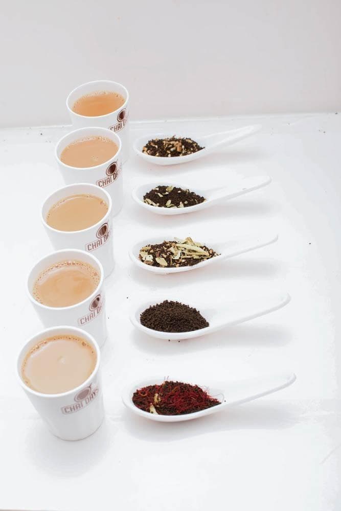 Five white Chai Days cups of milky tea in a row beside five white spoons, each holding a different dry tea or spice blend, on a white surface